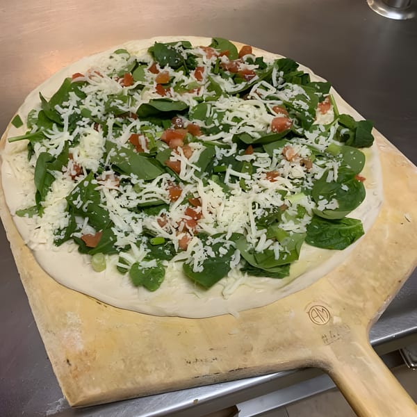 Unbaked Roma Ristorante Italiano pizza with spinach, diced tomatoes, and shredded cheese on a wooden peel.