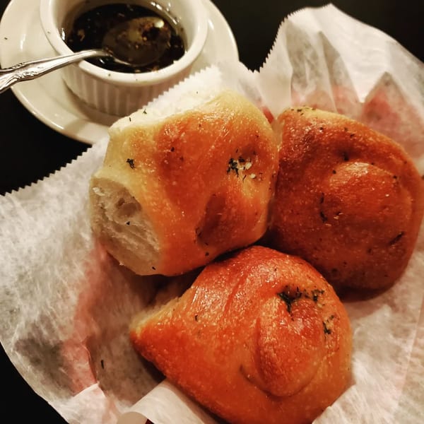 Three golden-brown Italian bread rolls with herbs in a paper-lined basket, next to a dish of dipping oil.